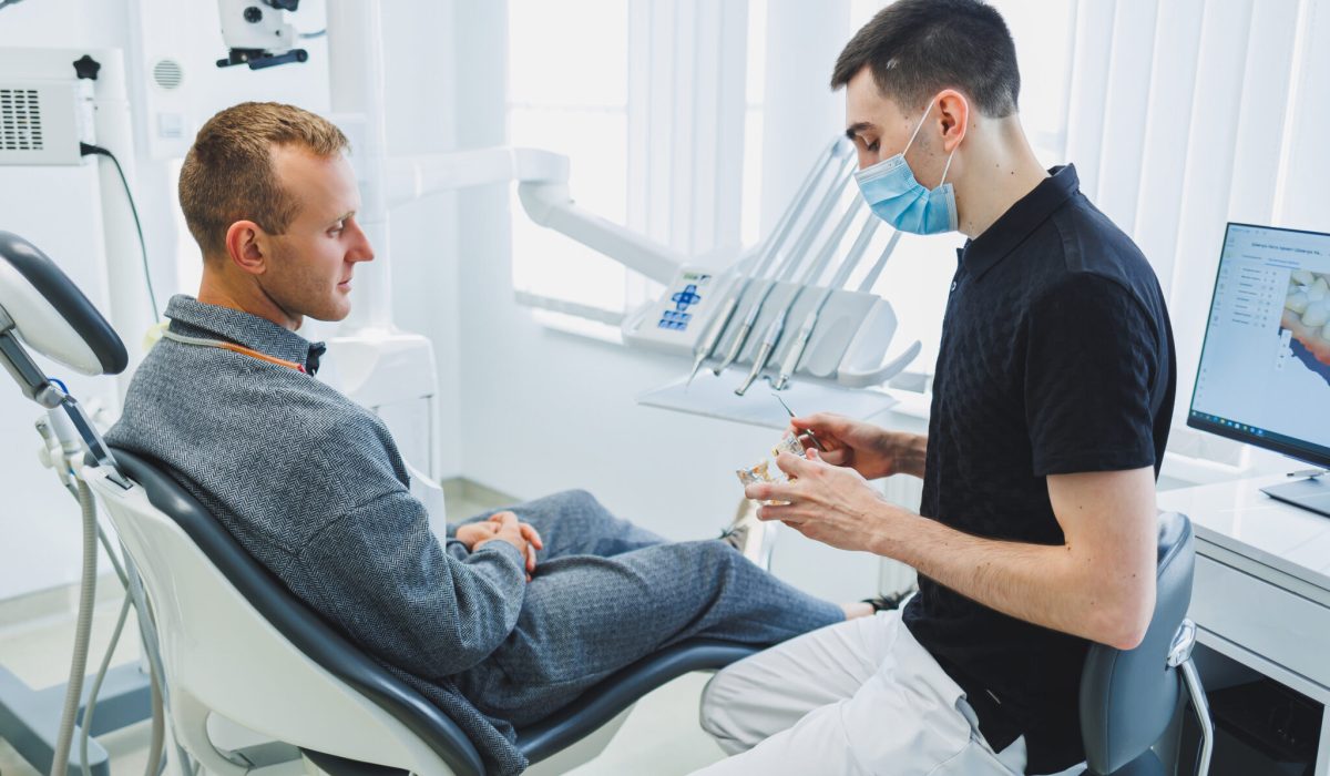 A dentist in a modern dental office tells a patient about dental care. A man sits in a dental chair and listens to a dentist.